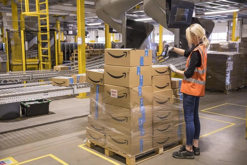 Warehouse worker in a safety vest scanning Amazon boxes stacked on a pallet next to conveyor belts inside an Amazon FBA fulfillment center