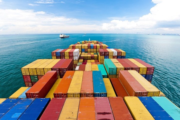 Vue du pont d'un porte-conteneurs entièrement chargé naviguant sur des eaux bleues et limpides, avec des conteneurs multicolores empilés en rangées bien ordonnées.