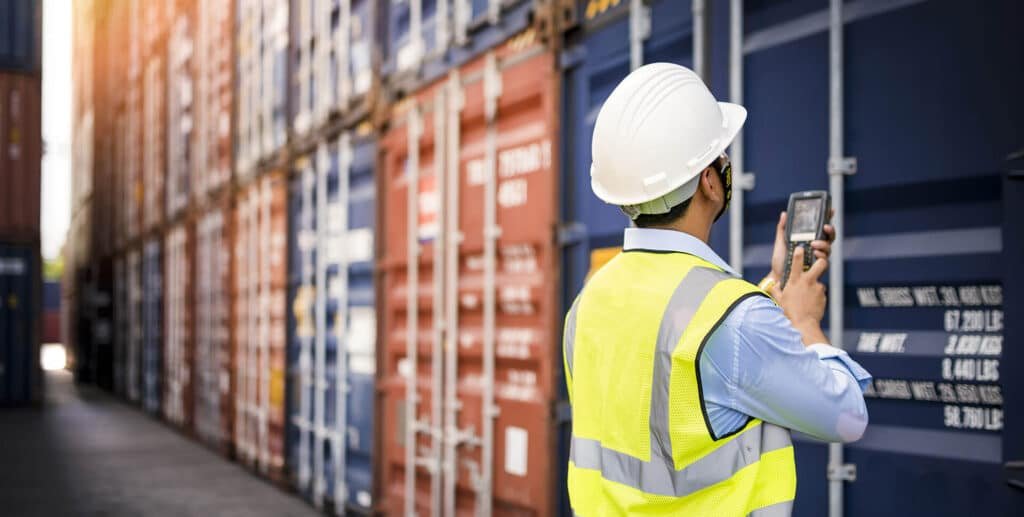 A worker in a high-visibility vest and hard hat using a handheld device to scan a container in a warehouse filled with stacked shipping containers, highlighting custom and insurance services.