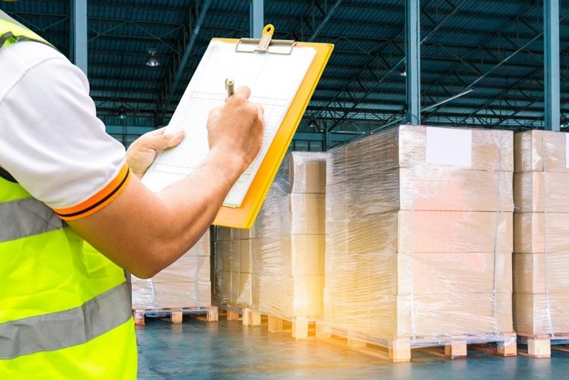 Warehouse worker in safety vest holding a clipboard and inspecting palletized boxes wrapped in plastic in a bonded warehouse