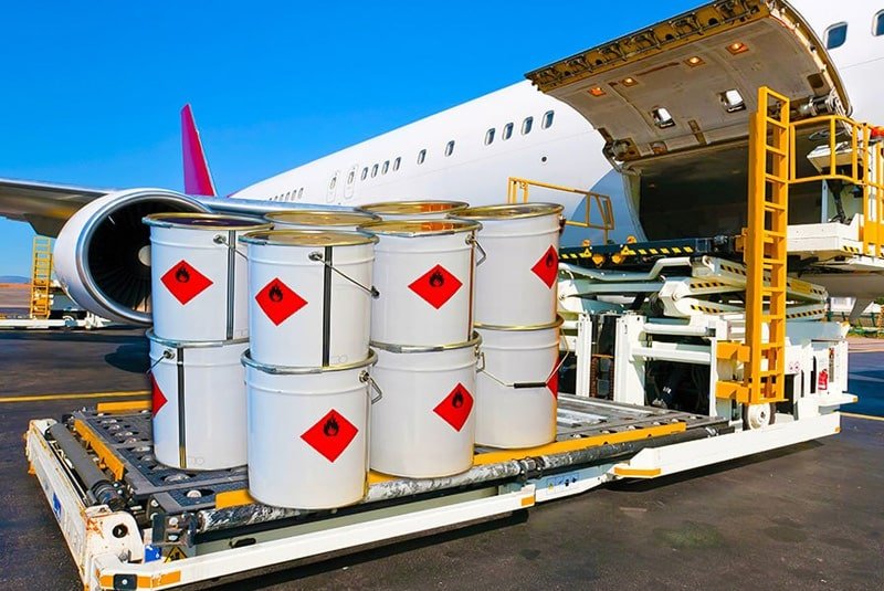 White barrels displaying red flammable hazard diamonds being loaded onto a cargo airplane via a loader