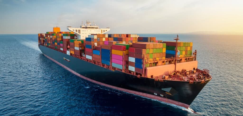 A large container ship loaded with multicolored cargo containers sailing across open ocean waters under a golden sunset sky
