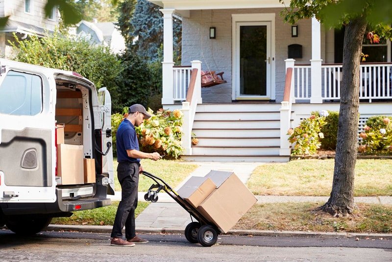 Delivery driver unloading boxes from a van onto a hand truck in front of a suburban home during door-to-door service