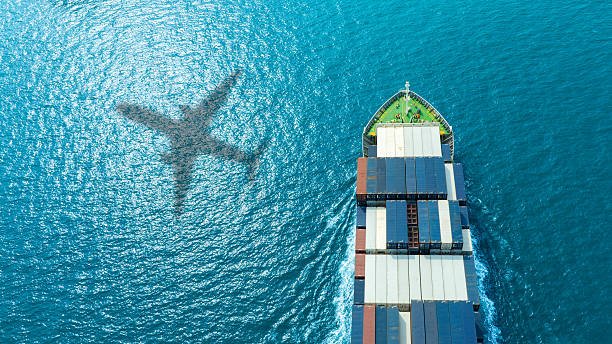 An aerial view of a cargo ship carrying containers on the ocean, with the shadow of a plane reflected on the water, symbolizing combined sea and air shipping.