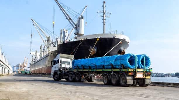 Flatbed truck carrying heavy rolled materials next to a docked freighter