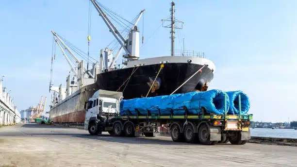 Flatbed truck carrying heavy rolled materials next to a docked freighter
