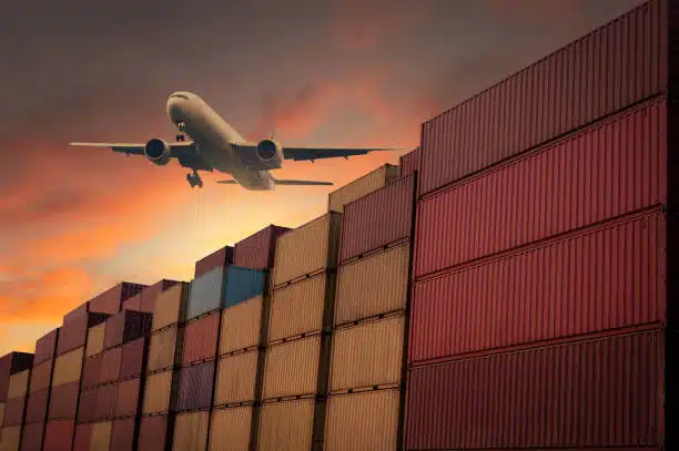 Airplane flying over stacked cargo containers during sunset, representing air freight from China to Venezuela