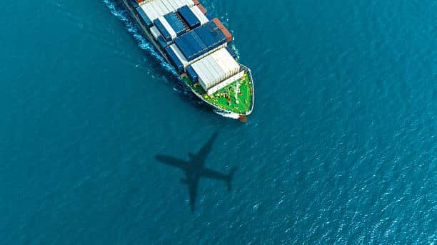 Aerial view of a cargo ship with airplane shadow on water, illustrating logistics from China to Hungary.