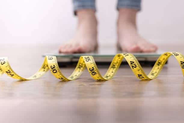 Feet on scale with measuring tape in foreground representing measurement of volume and weight