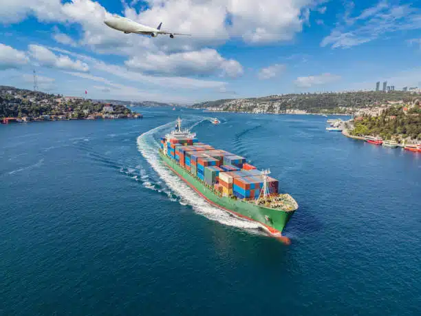 Cargo vessel navigating sea route toward Nigeria with airplane flying above