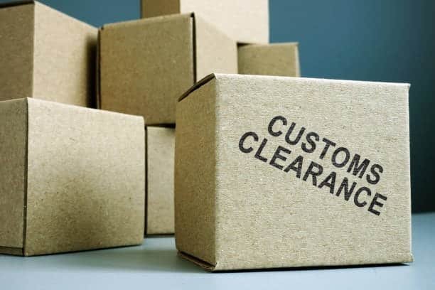 A group of brown cardboard boxes on a surface, with one box in the foreground stamped with the text "CUSTOMS CLEARANCE" in bold black letters.