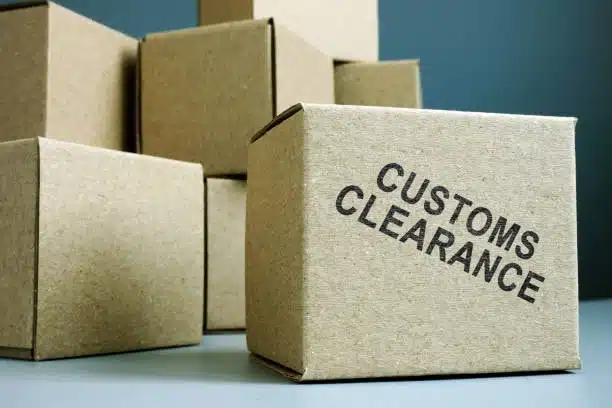 A group of brown cardboard boxes on a surface, with one box in the foreground stamped with the text "CUSTOMS CLEARANCE" in bold black letters.