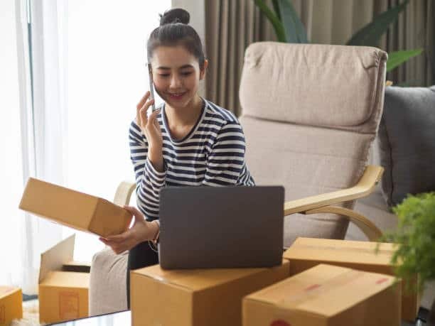 Woman working from home, managing dropshipping orders on a laptop while surrounded by parcels