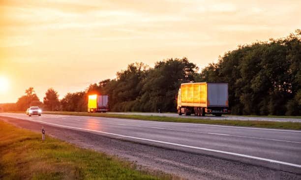 Two LTL freight trucks driving along a highway at sunset, delivering goods