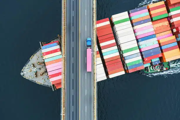 Top-down view of ocean freight container ship crossing under a highway bridge with truck passing overhead