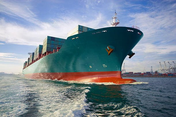 Large ocean freight container vessel approaching port with stacked shipping containers and port cranes in the background