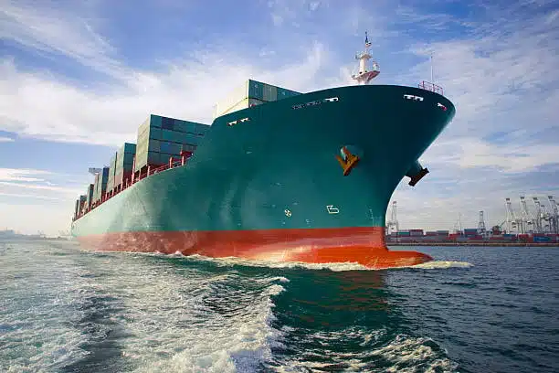 Large ocean freight container vessel approaching port with stacked shipping containers and port cranes in the background
