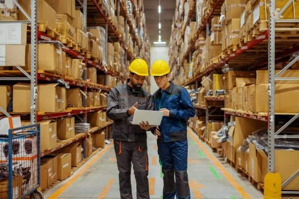 Two warehouse workers with hard hats reviewing documents among shelves stacked with parcels