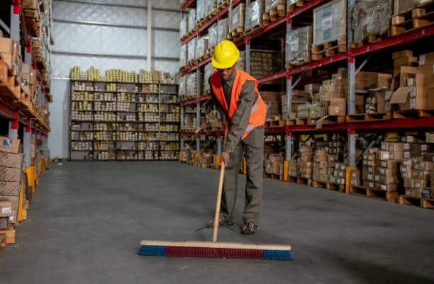 Worker in safety vest sweeping floor of warehouse filled with export cartons