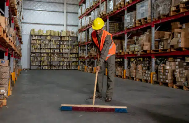 Worker in safety vest sweeping floor of warehouse filled with export cartons