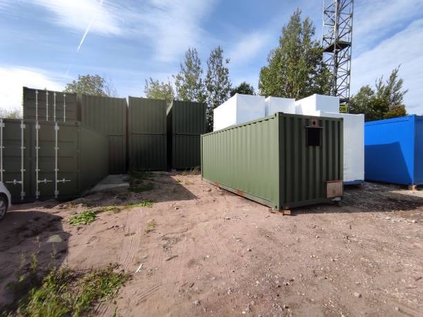 A green 20 feet container stationed in an outdoor logistics yard, with other stacked storage units visible in the background.